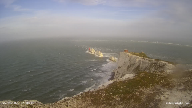 A wide view of a coastal scene featuring steep white chalk cliffs with grassy tops, a building located on the edge of the cliffs, and a lighthouse on a rock formation offshore in the distance. The ocean waves are crashing against the cliffs and rocks, with a partly cloudy sky overhead.