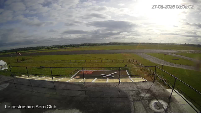 View of an outdoor airfield taken from a webcam at Leicestershire Aero Club. The image shows a grassy area surrounding a small runway, with some fencing and a windsock on the left side. The sky is partly cloudy with the sun shining through, and the time and date are displayed in the top right corner as 27-05-2025 08:00:02.