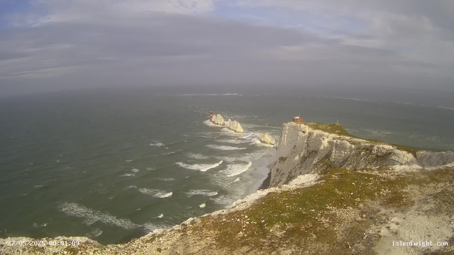 A wide view of white chalk cliffs overlooking the ocean, with a lighthouse and a small building on the edge of the cliff. The sea has gentle waves, and the sky is mostly cloudy with some patches of sunlight. There are large rock formations in the water near the base of the cliffs.