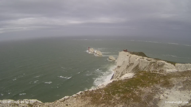 A view of dramatic white chalk cliffs and rocky formations along the coastline on a cloudy day. The sea is choppy with small waves, and there is a path or railing along the edge of the cliffs. A small building or shelter is visible on top of the cliffs. The scene is overcast with dark clouds, and the ocean extends to the horizon in the distance.