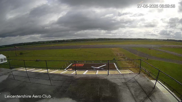 A view from a webcam at Leicesterhire Aero Club showing a cloudy sky over an open airfield. In the foreground, there's a helipad with a large white helicopter silhouette painted on it. A flagpole with a flag is visible on the left side. The grassy field extends into the distance, with runways and taxiways visible on the right side. The timestamp indicates the photo was taken on May 27, 2025, at 8:59 AM.