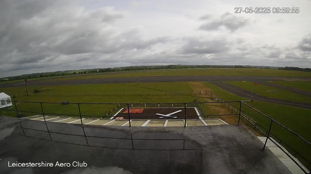 A view of an airfield on a cloudy day, with a helicopter landing pad in the foreground. The pad has a white helicopter silhouette painted on it, surrounded by a white border and a small red rectangle. The airfield extends into grassy areas, with a few small structures and signage visible. The background shows a long, straight runway, with trees and fields beyond. The sky is filled with dark, cloudy clouds. The timestamp in the upper right corner reads "27-05-2025 09:59:55", and the bottom left corner has the text "Leicestershire Aero Club".