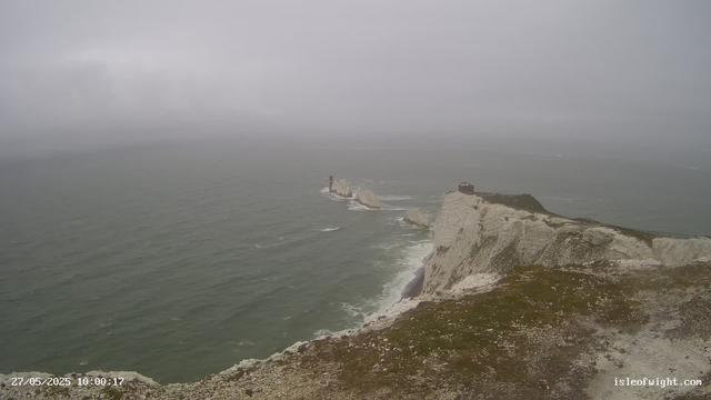 A panoramic view of the white chalk cliffs along a misty, overcast shoreline. The cliffs rise sharply from the sea, with a small observation building near the edge. The ocean appears choppy, with waves crashing against the base of the cliffs. In the distance, a lighthouse is visible further out at sea. The sky is filled with thick gray clouds, creating a moody atmosphere. The image is timestamped "27/05/2025 10:00:17" and has "isleofwight.com" written in the bottom right corner.