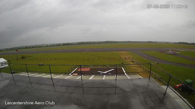 A wide view of an airfield on a cloudy day. In the foreground, there is a rooftop with a railing, a small helipad with a large white "H" and some markings, and a tractor parked on the right. Below the balcony, there's a grassy area with a flagpole. Across the airfield, there are taxiways and a small airplane parked on the grass. The background features open fields with a line of trees far away under an overcast sky. The timestamp indicates the date and time as 27th May 2025 at 11:00 AM. The image is labeled "Leicestershire Aero Club."