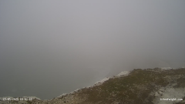 A foggy, overcast landscape with a rocky, uneven surface in the foreground. The sky is thick with fog, obscuring most of the background and creating a grey, misty atmosphere. The ground appears dark with patches of white and light-colored rocks or snow along the edge. The image has a timestamp in the lower left corner showing "27/05/2025 11:01:01" and a URL "isleofwight.com" in the lower right corner.