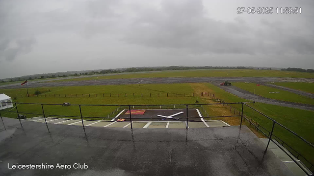 A view from a webcam at Leicesterhire Aero Club on a cloudy day. The image shows a rooftop with a clock embedded in the surface, displaying the time as approximately 11:59.51. Beyond the rooftop, there is a grassy area enclosed by a fence, a windsock on a pole, and a small paved area with a black helicopter silhouette painted on it. In the background, there are taxiways with a small vehicle moving across, and the open sky with gray clouds covering the scene. The date and time in the top right corner indicate it was taken on May 27, 2025, at 11:59:51.