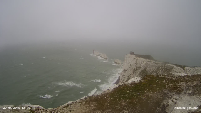 A foggy seascape featuring white chalk cliffs extending into the ocean. The cliffs have dark patches of vegetation on the top, and a winding path runs along the edge. The ocean below is rough with white-capped waves, and the sky is overcast with dense fog obscuring the horizon. A small building is visible on the top of the closest cliff, and a lighthouse can be seen further along the shoreline in the distance. The image is timestamped "27/05/2025 12:00:46" and is from islefowight.com.