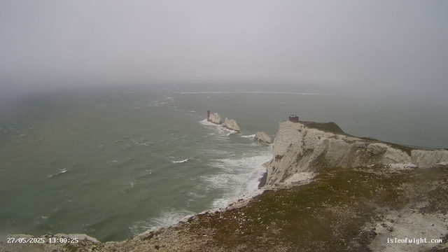 A view from a webcam showing a foggy coastal scene with white chalk cliffs and a small building at the top. The cliffs overlook a choppy greenish-grey sea, with a narrow pathway running along the edge of the cliffs. Some rock formations with a lighthouse are visible in the water near the cliffs. The image is timestamped "27/05/2025 13:00:25" and has the website "isleofwight.com" in the bottom right corner.
