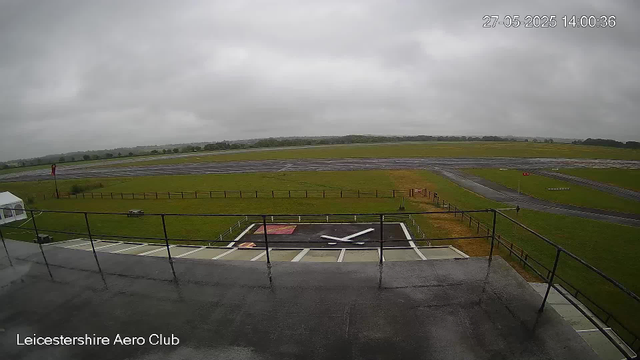 A view from a webcam at Leicesterhire Aero Club shows a cloudy, overcast sky above a grassy airfield with runways stretching into the distance. In the foreground, there is a helipad with a white helicopter silhouette, and a small paved platform with a railing. To the left, a white tent is visible, and a flag pole with a red flag is near the tent. The landscape is mostly flat with some distant trees and rolling hills under a gray sky. The timestamp in the top right corner reads "27-05-2025 14:00:36".