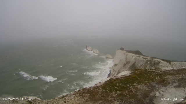 A view of a foggy coastline with white chalk cliffs and a walking path along the top. The sea below is rough with visible waves crashing against the rocks at the base of the cliffs. In the distance, a small structure is visible on the cliff edge, and there are several large rocks in the water near the shoreline. The scene has a grey, overcast sky, and the overall atmosphere appears cold and misty.