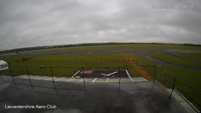 An overcast sky with gray clouds above an airfield at Leicestershire Aero Club. In the foreground, there's a rooftop with a railing, and a helicopter landing pad marked with a large white formation on a black surface. The grassy area around the runway is fenced, and there is a small bench near the fence. Multiple runways extend into the distance across the landscape.