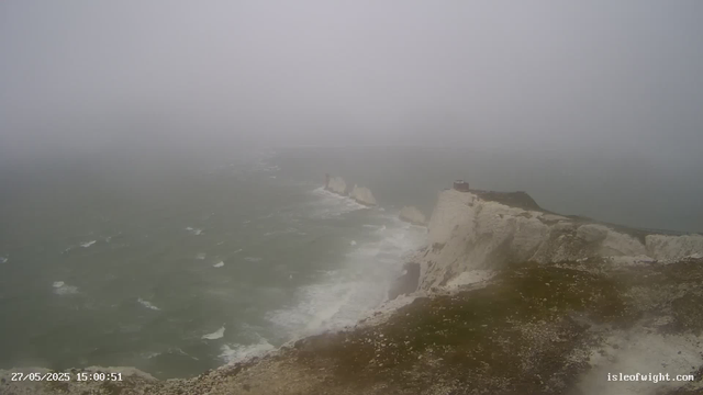 A foggy scene of white chalk cliffs along a rugged coastline with the sea crashing against the rocks below. A small building is visible on top of the cliffs, and the overall atmosphere is misty and damp.