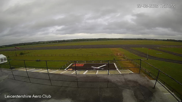 A wide view of an outdoor airfield on a cloudy day, with a large clock painted on the ground in the foreground, showing the time as 10:15. There is a flagpole with a flag, a small white tent on the left, and green grassy areas bordering the tarmac. The background features open fields and a low horizon under a gray, overcast sky. The top right corner displays a timestamp of "27-05-2025 17:00:55," and the bottom left corner reads "Leicestershire Aero Club."