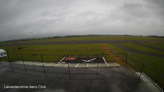 An overcast sky above a flat, grassy airfield with a runway stretching into the distance. In the foreground, there is a helipad with a large white "H" and a small red rectangular marking on the ground. A black metal railing surrounds the area, and a flagpole with a flag is visible on the left side. The words "Leicestershire Aero Club" are displayed in the bottom left corner. The scene appears to be calm with no visible aircraft or people.