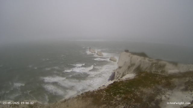 A foggy scene of white chalk cliffs along a choppy, gray-green sea. Waves crash against the base of the cliffs, partially obscuring the view. There are several prominent chalk stacks standing in the water further out, with one in the distance near the horizon. The sky is overcast, and visibility is limited due to the fog. A timestamp and a website URL are visible in the bottom corners.