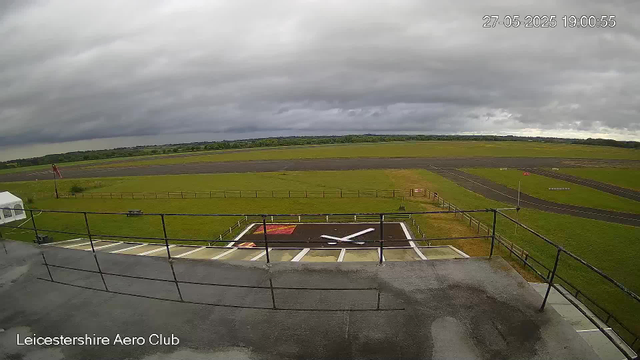 An outdoor view of Leicestershire Aero Club showing a cloudy sky over an empty runway with green grass on either side. In the foreground, there is a raised platform with metal railings and a large helicopter rotor hub marked on the tarmac. There is also a small white tent on the left and a flagpole with a flag on the left side of the image. The timestamp indicates the date and time as 27-05-2025 at 19:00:55.