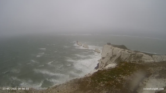 A foggy view of white chalk cliffs along a rugged coast, with the sea crashing below. The sky and horizon are obscured by dense fog, creating a mysterious and muted atmosphere. A small structure is visible at the top of the highest cliff on the right. The image has a timestamp of "27/05/2025 20:00:01" and the website "isleofwight.com" is displayed in the bottom right corner.