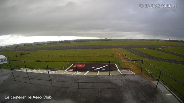 The image shows an outdoor scene at Leicesterhire Aero Club with a cloudy sky overhead. In the foreground, there is a small helipad marked with a large white "H" and red and orange markings nearby. Surrounding the helipad is a short black railing. To the left, there is a white tent, and a flagpole with a red flag flying. The background consists of expansive grassy fields and a paved track or runway stretching into the distance under a dark, overcast sky. The timestamp indicates the photo was taken on May 27, 2025, at 8:00 PM.