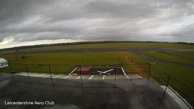 A view from a webcam shows an overcast sky with dark, heavy clouds above an airfield. In the foreground, there is a rooftop with a railing and a large helipad featuring a prominent white helicopter symbol painted on it. To the left, a small tent is set up near a windsock, which is pointing slightly downward. Beyond the helipad, a grassy area demarcates the runway and taxiways, which extend into the distance. The landscape around the airfield is flat with some distant trees and low hills on the horizon. The timestamp indicates the date and time as 27-05-2025 at 21:00:20. The bottom left corner displays the text "Leicestershire Aero Club."