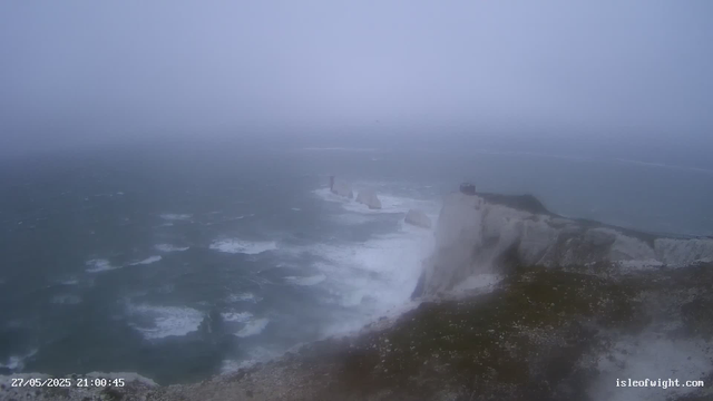 A view of a rugged coastline with tall white cliffs and crashing waves below, under a foggy, overcast sky. The scene is shrouded in mist, limiting visibility of the distant horizon.
