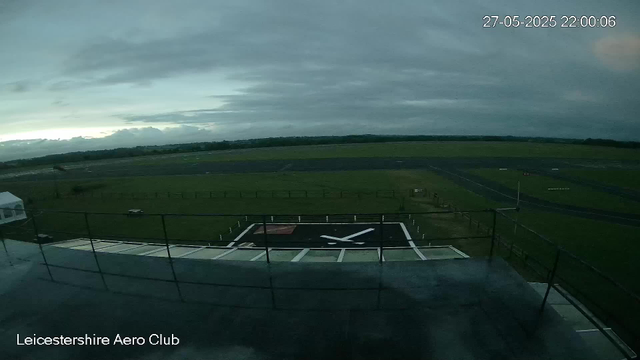 A view from a webcam at Leicesterhire Aero Club shows a green open field with a runway, a black helipad with a white helicopter symbol, and a cloudy sky overhead. The timestamp reads 27-05-2025 22:00:06.