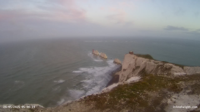 A panoramic view of white chalk cliffs rising from the sea, with a small building on top of the nearest cliff. The coastline extends into the distance with rock formations and a lighthouse visible on one of the rocks in the water. The sky is partly cloudy with soft pastel colors, indicating early morning or late evening. The timestamp in the bottom left corner shows "28/05/2025 05:00:14" and the website "isleofwight.com" is shown in the bottom right corner.