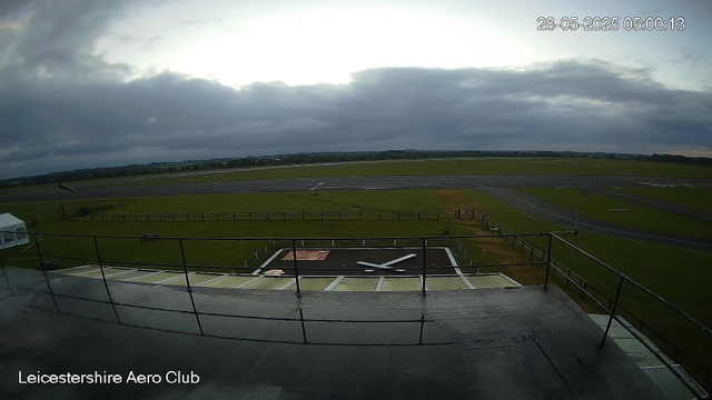 An overcast sky with dark clouds over an empty airfield at Leicester Aero Club. The runway and taxiways are visible in the distance, with a grassy area in between. In the foreground, there is a rooftop area with a rail, and a helipad with a large white "H" symbol. A windsock is near the left side of the image, and a small tent or shelter is visible on the left. The scene is calm, and the timestamp shows the date and time as 28-05-2025, 05:00:13.