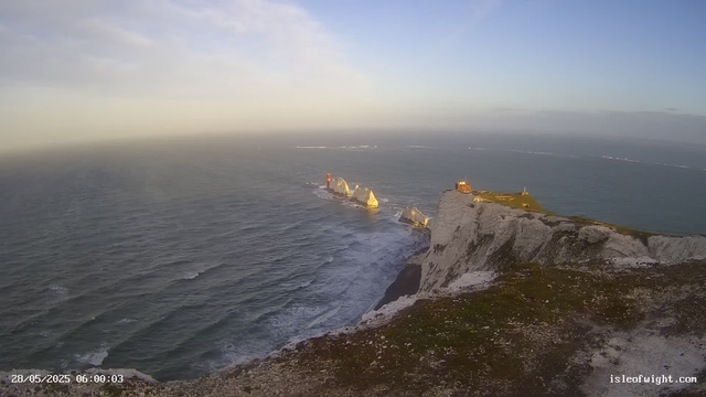 An aerial view of white chalk cliffs with a grassy top coastline, extending into the ocean. There are several white rock formations and a small lighthouse on the cliffs. The ocean waves are gently crashing against the rocks below, and the sky is clear with a soft gradient from light to darker blue. The scene is illuminated by bright sunlight, and a timestamp at the bottom left reads "28/05/2025 06:00:03".