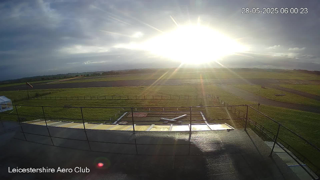An early morning view at Leicesterhire Aero Club with the sun rising on the horizon, casting bright light across the open grassy area and surrounding airstrip. The sky is partly cloudy with dark clouds on the left and clear skies on the right. The scene is viewed from an elevated platform with metal railings in the foreground. The date and time are displayed in the top right corner: 28-05-2025, 06:00:23.