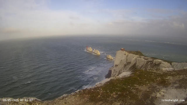 A view of rugged white chalk cliffs overlooking the ocean. In the distance, there are several large sea stacks rising from the water, with a small red-and-white lighthouse situated on the tallest stack. To the right, a building is perched on the edge of the cliff. The sky is partly cloudy, and the time stamp shows the date as 28/05/2025 at 07:01.