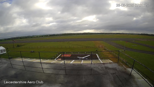 An outdoor view from a webcam at Leicesterhire Aero Club shows a cloudy sky over a large open grassy area and a runway in the distance. In the foreground, there is a rooftop with a black helipad marked with a white "H" and a red square adjacent to it. A clock and calendar date are visible in the upper right corner, showing "28-05-2025 07:01:05". The bottom left corner displays the location, "Leicestershire Aero Club."