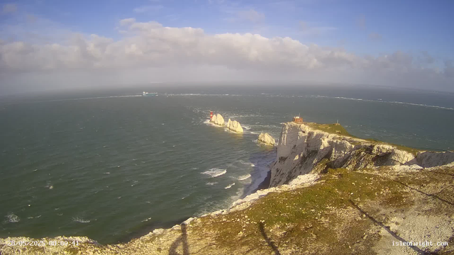 Aerial view of white chalk cliffs along the coastline, with a grassy area and a small building on top. The cliffs extend into the sea, with waves crashing at the base. A lighthouse stands near the edge of the cliffs. In the background, a ship and a distant wake in the water are visible under a partly cloudy sky.
