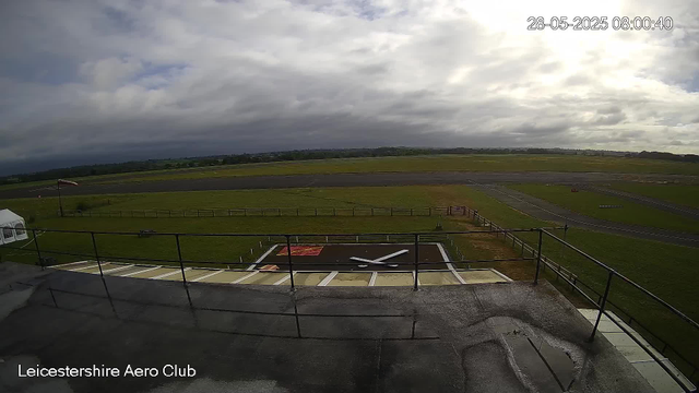 A wide view of an airplane runway and grassy fields under a cloudy sky, taken from a webcam at Leicestershire Aero Club. The time and date are displayed in the top right corner as 28-05-2025 08:00:40. There is a large clock face on the ground in front of the image, with the clock's hands pointing to around 4:00.