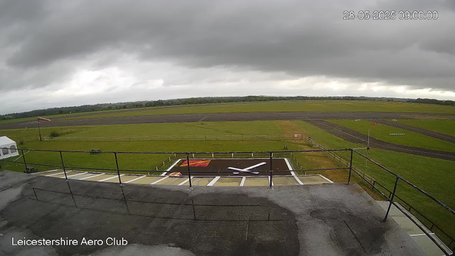 An outdoor view from a webcam at Leicester Aero Club showing a cloudy sky over an open grassy area and a small runway. In the foreground, there is a helipad with a large white helicopter marking, surrounded by a safety railing. To the left, there is a small white tent and a flagpole with a flag. The scene is overcast, with dark clouds covering most of the sky. The timestamp indicates the photo was taken on May 28, 2025, at 9:00 AM.