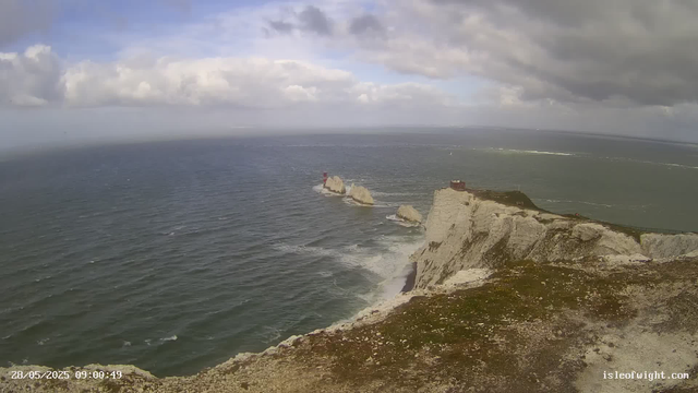 A wide view of the ocean with white chalk cliffs in the foreground, a lighthouse on one of the smaller rocks offshore, and a small building atop the main cliff. The sky is partly cloudy with patches of blue, and the water appears calm with gentle waves.