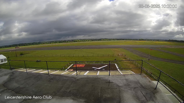 A View from a Webcam at Leicester Aero Club showing a cloudy sky over a runway and grassy fields. In the foreground, there is a helipad with a white helicopter silhouette marking, a small flag, and a railing. The date and time stamp on the top right corner reads "28-05-2025 10:00:00." The weather appears overcast with dark clouds, and the overall scene is calm and empty of people or aircraft in motion.