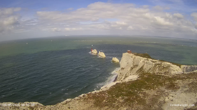 A view of a coastal scene with white chalk cliffs, a lighthouse on a small rocky outcrop in the sea, and a building on top of the cliff. The ocean stretches out with sailboats visible in the distance, under a partly cloudy sky. The image is timestamped "28/05/2025 10:01:08" and has the website "isleofwight.com" in the lower right corner.