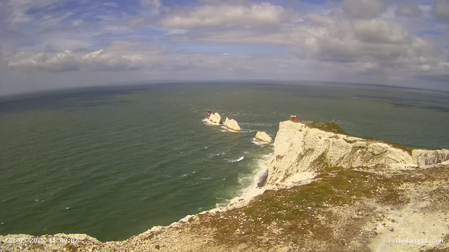 A view of white chalk cliffs along the coast with waves crashing at their base. There is a small rocky islet and a lighthouse in the distance, and a building on top of the cliff to the right. The sky is partly cloudy with patches of blue.