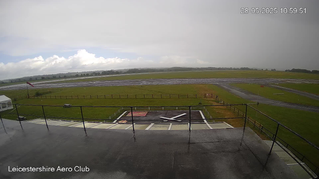 An aerial view of Leicestershire Aero Club taken from a webcam. The image shows a grassy airfield with taxiways and runways extending into the distance under a cloudy sky. In the foreground, there is a helipad with a large X mark, surrounded by a chain-link fence, and a small white tent on the left. A windsock is visible near the fence, and the landscape beyond the airfield is flat with some trees on the horizon. The timestamp on the image indicates it was captured on 28th May 2025 at 10:59:51.