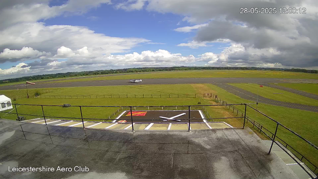 A view from a webcam at Leicestershire Aero Club shows a cloudy sky over an airfield with a small airplane taxiing on the runway in the distance. In the foreground, there is a helipad with a large white "H" marking, surrounded by a black railing, and a small white tent on the left side. The grassy areas are well-maintained, and the sky is partly cloudy with patches of blue visible. The timestamp in the top right corner reads "28-05-2025 12:00:12".