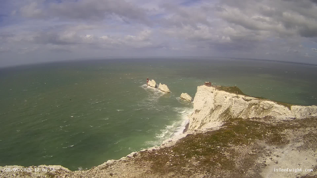 A view of the white chalk cliffs along the coast with a lighthouse and a small building on top, extending into a choppy greenish sea under a cloudy sky.