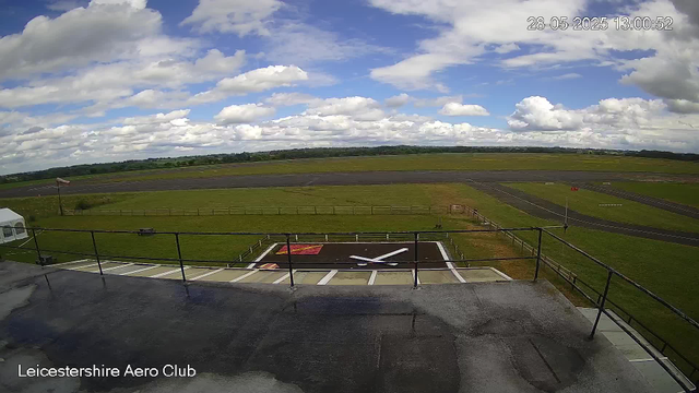 An outdoor view from a webcam at Leicestershire Aero Club shows a bright sky with scattered white clouds and a flat green landscape. In the foreground, there is a small helicopter pad marked with a large white helicopter rotor symbol, surrounded by a black safety fence. To the left, a white tent or temporary structure is visible, and a flagpole with a flag is near the tent. The runway and taxiways extend into the distance, bordered by grassy areas and some small objects or equipment. The timestamp indicates the date and time as 28th May 2025 at 13:00.