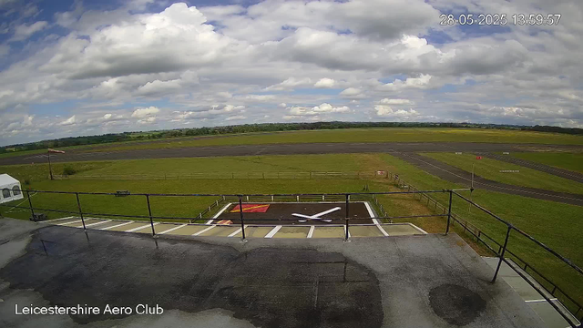 A wide view of an airfield shows a runway stretching across a grassy landscape with a cloudy sky overhead. In the foreground, a rooftop helipad marked with a large white helicopter symbol and bordered by a metal railing is visible. To the left, part of a small white tent or building is seen, and a windsock is positioned at the edge of the grassy area. The bottom of the image has the text "Leicestershire Aero Club." The top right corner displays the date and time: 28-05-2025 13:59:57.
