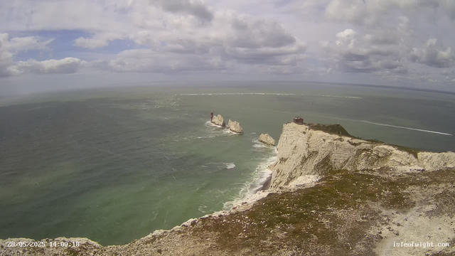 A view of white chalk cliffs with a grassy top, overlooking the ocean. There are three large rock formations with one having a lighthouse on top, located in the water near the cliffs. The sky is partly cloudy with patches of blue. The ocean waves are gently crashing against the rocks and coastline. The perspective appears to be from a webcam mounted on the cliff edge.