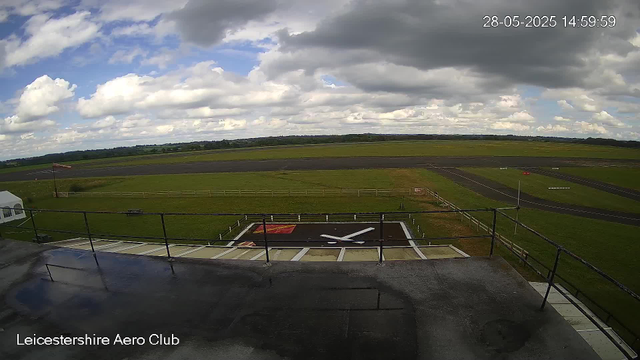 A view from a webcam at Leicestershire Aero Club shows an open grassy airfield under a partly cloudy sky. In the foreground, there is a large clock mounted on a platform, with white hour and minute hands. The timestamp in the top right corner indicates the date as May 28, 2025, and the time as 14:59:59. The horizon reveals a vast expanse of green fields and distant trees. The scene appears calm with no aircraft or people visible.