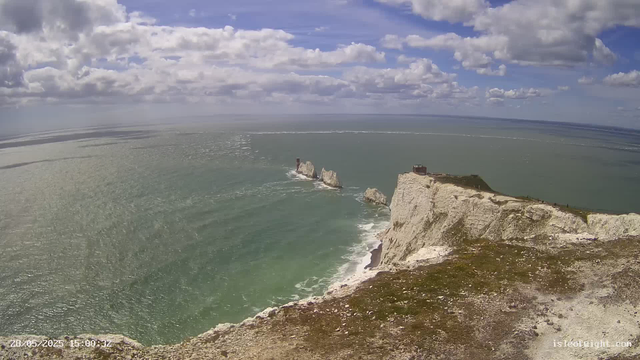 Aerial view of white chalk cliffs overlooking the sea with rock formations in the water, partly cloudy sky with blue patches, and a building on top of the cliff. The ocean is a greenish hue, and the coastline extends into the distance.