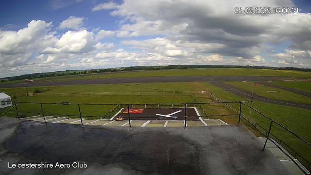 A view from a webcam at Leicestershire Aero Club shows a small aircraft parked on a tarmac area with a large runway in the background. The sky is partly cloudy, with a mix of white and gray clouds, and the landscape is primarily green fields. The date and time displayed are May 28, 2025, at 16:00:31.