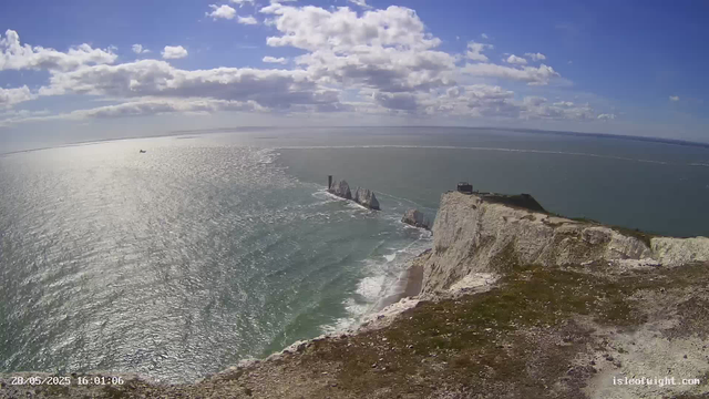 An aerial view of a coastal landscape featuring white chalk cliffs overlooking the ocean. The sky is partly cloudy with sunlight reflecting off the water. A small boat is visible far in the distance on the sea. There is a structure on top of the cliffs, with a few patches of greenery and a pathway leading along the edge.