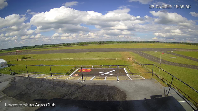 A view from a webcam overlooking an airfield with a large clock painted on the ground in the foreground, showing the time as 4:00. The sky is partly cloudy with white fluffy clouds and patches of blue sky. There is a flagpole with a waving flag on the left side, and a small tent can be seen near the flagpole. The airfield extends into grassy fields and taxiways, with some distant trees and landscape on the horizon. The image has a timestamp in the top right corner displaying the date and time as 28-05-2025 16:59:50. The bottom left corner has text that reads "Leicestershire Aero Club."