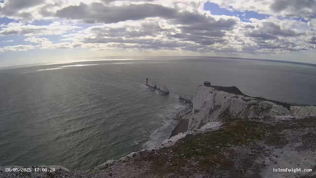 A view from atop white chalk cliffs overlooking the ocean, with several large rock formations or stacks rising from the water near the base of the cliffs. The sky is partly cloudy with patches of sunlight breaking through the clouds, and the sea appears calm with gentle waves. A small lighthouse or navigation tower is visible on the cliff edge. The date and time stamp in the bottom left corner reads "28/05/2025 17:00:20" and a website URL "isleofwight.com" is shown in the bottom right corner.